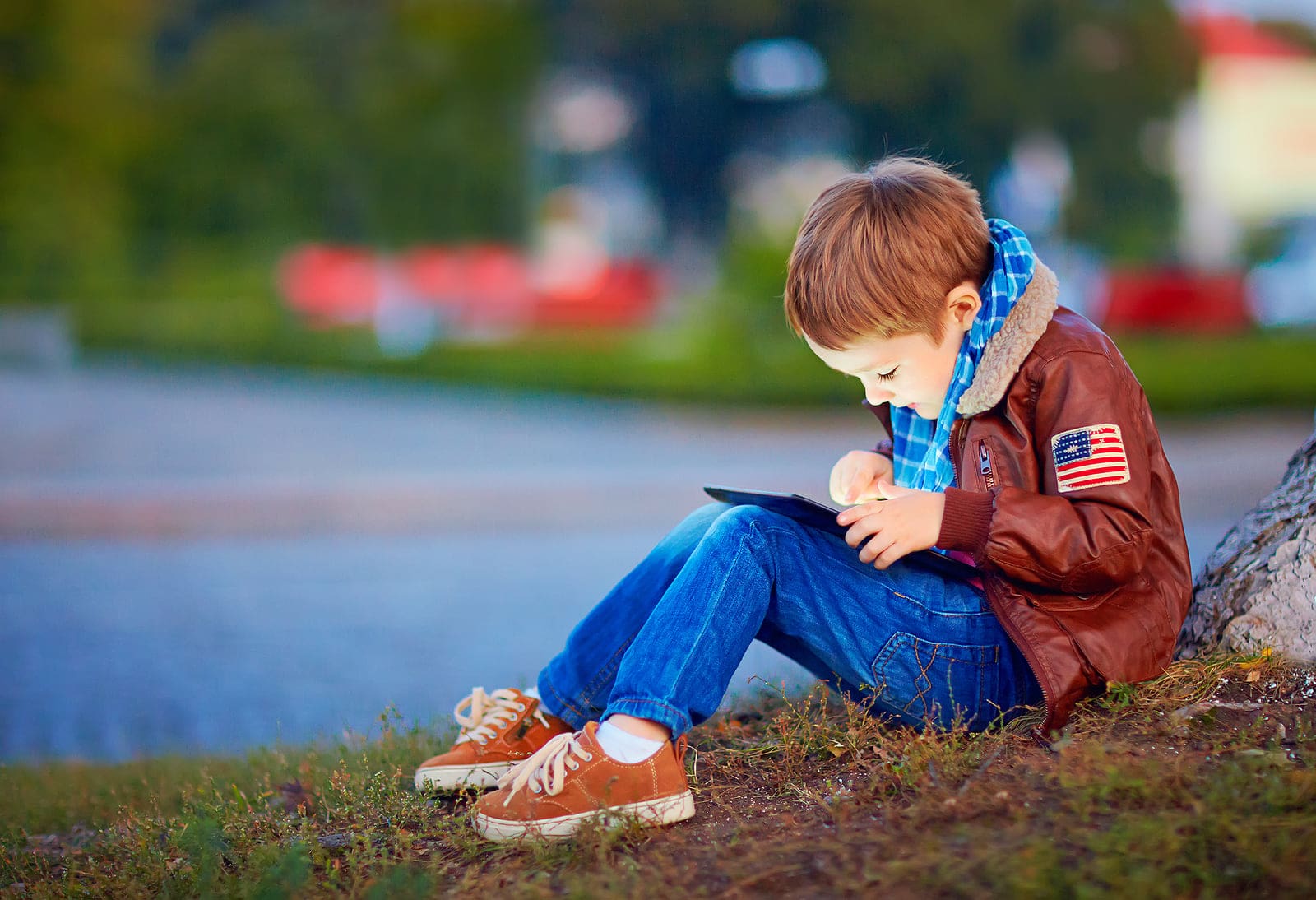 Child engrossed with tablet, wearing brown jacket, blue jeans, outdoor photo.