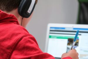 Person in red hoodie using laptop, wearing headphones, holding blue pen.