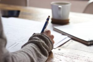 Person writing with blue pen on paper, wooden table, photo style.