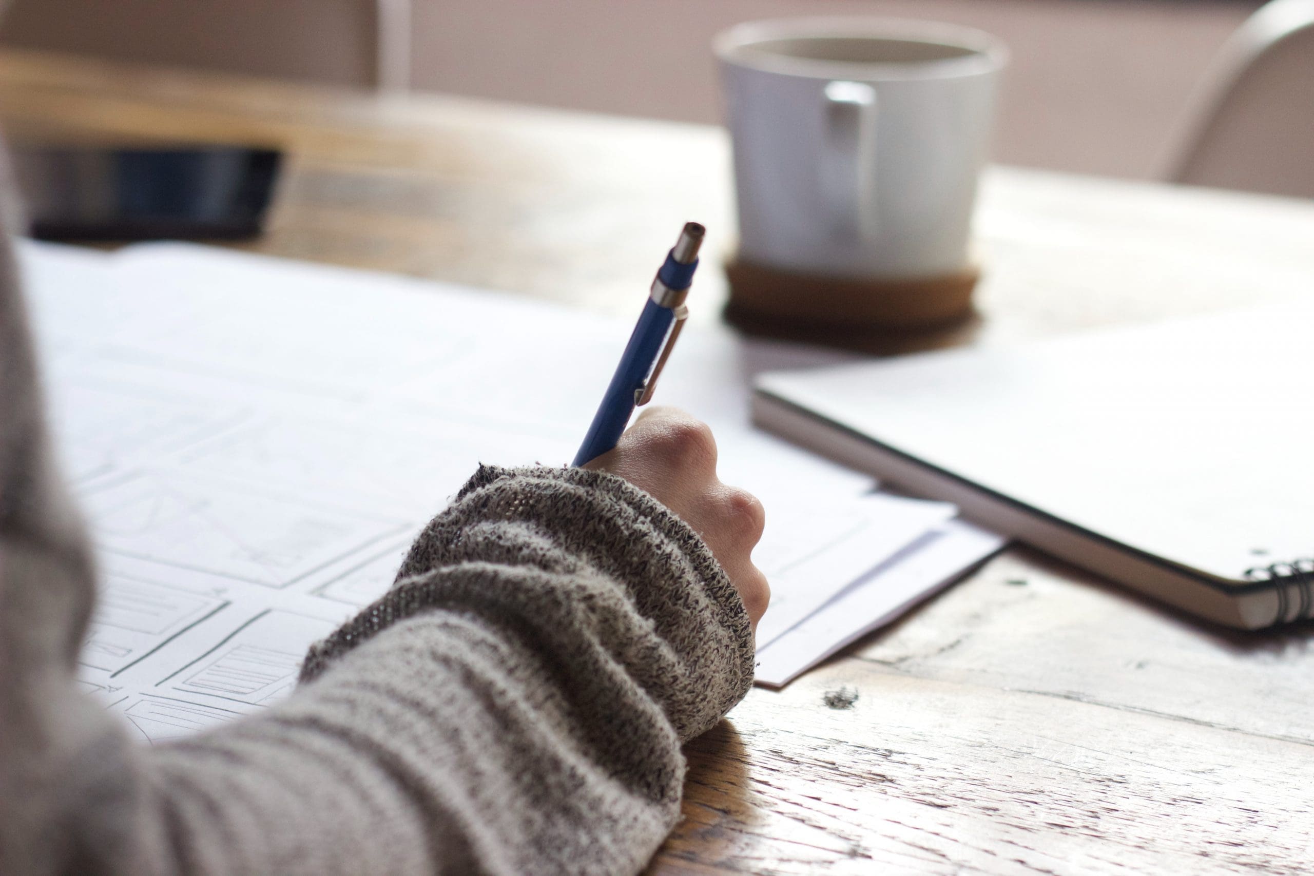 Person writing with blue pen on paper, wooden table, photo style.